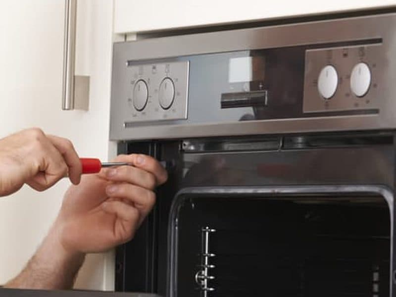 Close up of a built-in oven in a fitted kitchen, with a hand undoing a screw on the door frame.